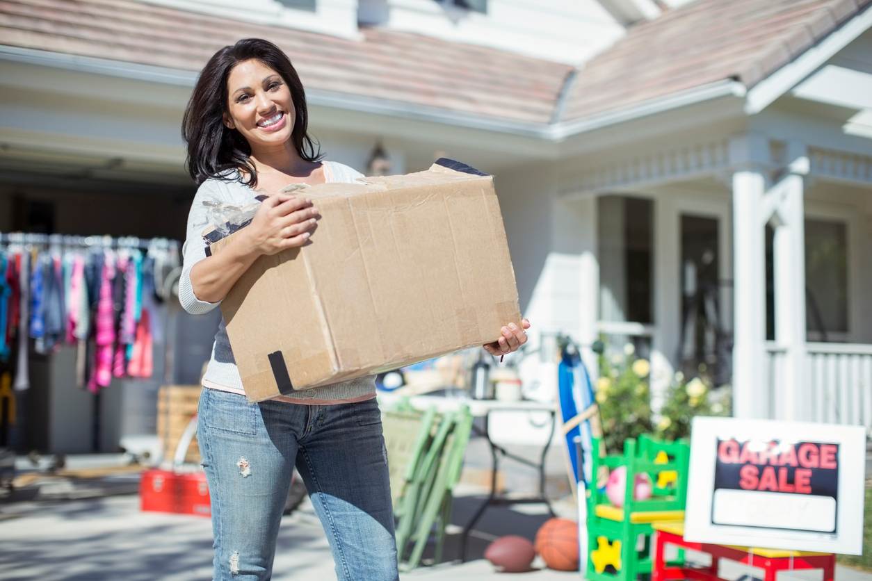 A smiling woman carries a cardboard box in front of a garage sale